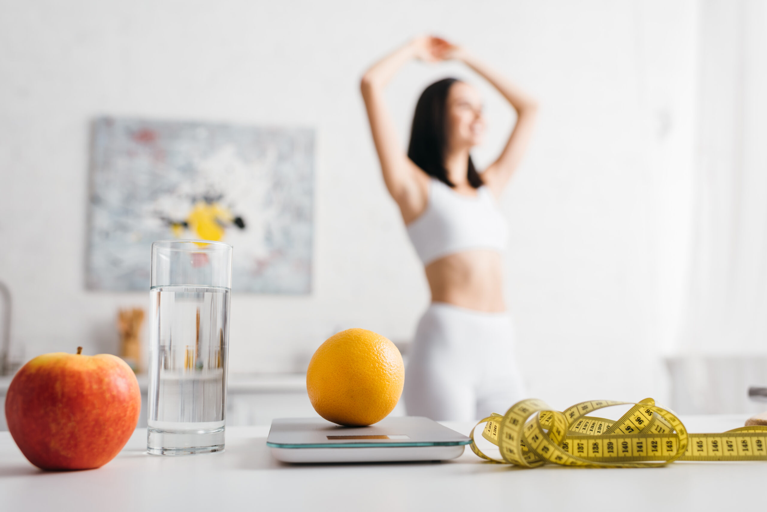 Selective focus of measuring tape, fruits with scales and glass of water on table and sportswoman in
